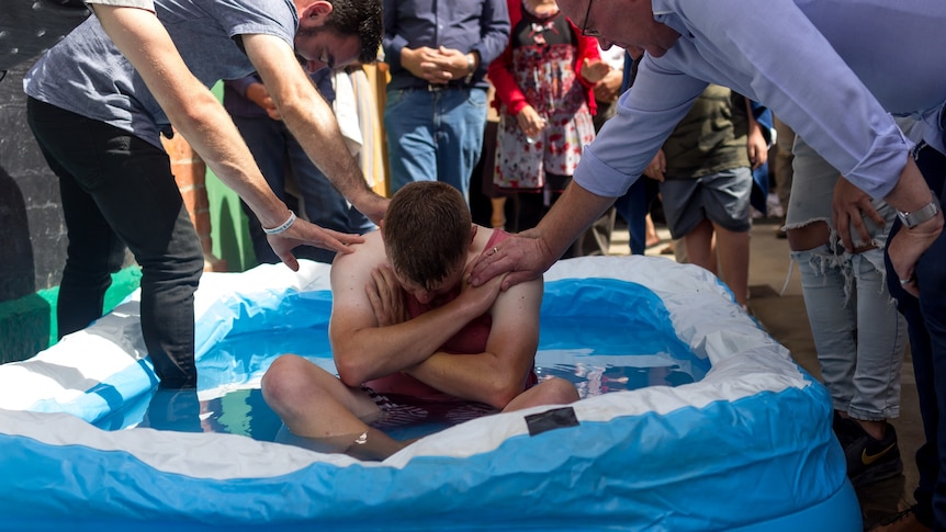 A group of people gathered around a man sitting in an inflatable pool.