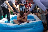 A group of people gathered around a man sitting in an inflatable pool.