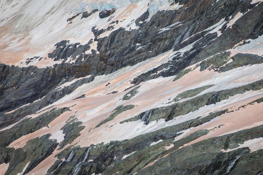 A close-up of a glacier shows sparse ice, tinged with red, covering black rocks.