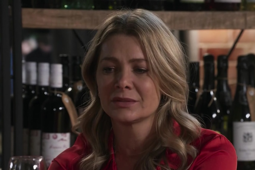 a woman in a red shirt looks sad. there are wine bottles on a shelf behind her.