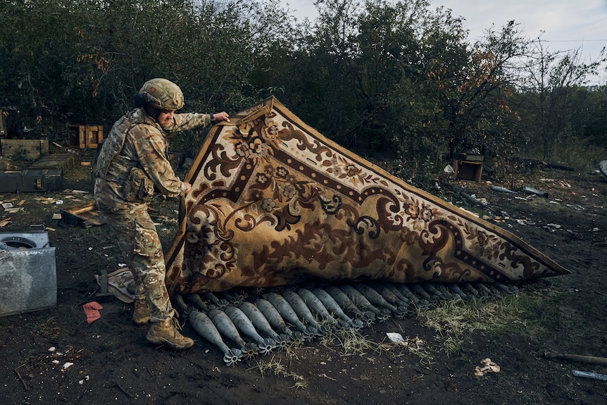 Un soldat en uniforme décolle un grand tapis du sol, révélant des munitions posées dans la boue