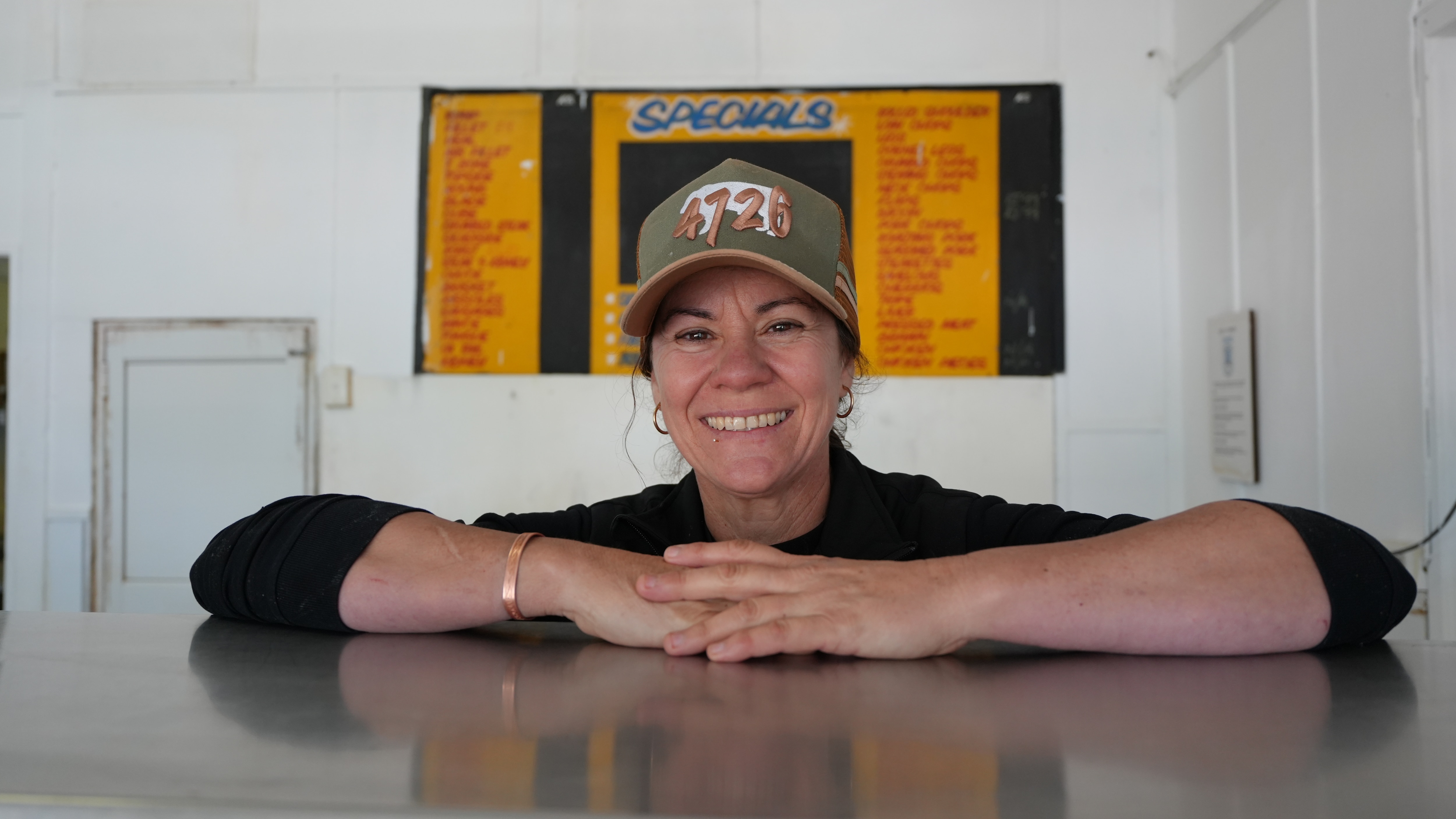 Woman standing behind counter at outback butcher shop smiling with her arms on the bench. She wears a black jumper and cap.