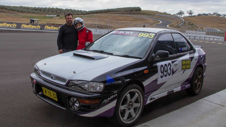 A man and a girl stand behind a car with a race circuit in the background
