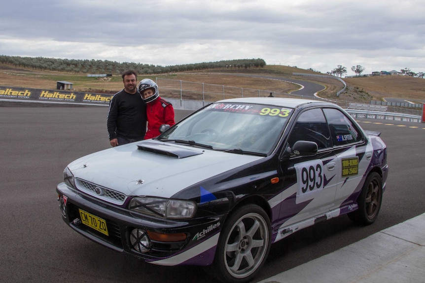 A man and a girl stand behind a car with a race circuit in the background