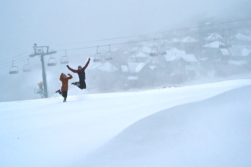 Two people jump happily in front of ski lifts as snow falls.