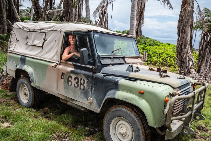 A woman pictured smiling in a camouflage truck in a tropical island looking forest setting.