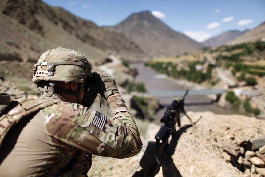 Soldier is seen from behind as he surveys landscape through binoculars.