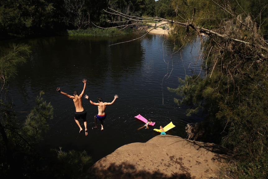 Two young people jump from a large raised rock into dark water below, with their arms flailing above their heads.