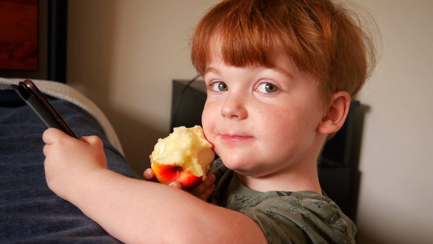 Headshot of a young boy holding a phine and eating an apple.