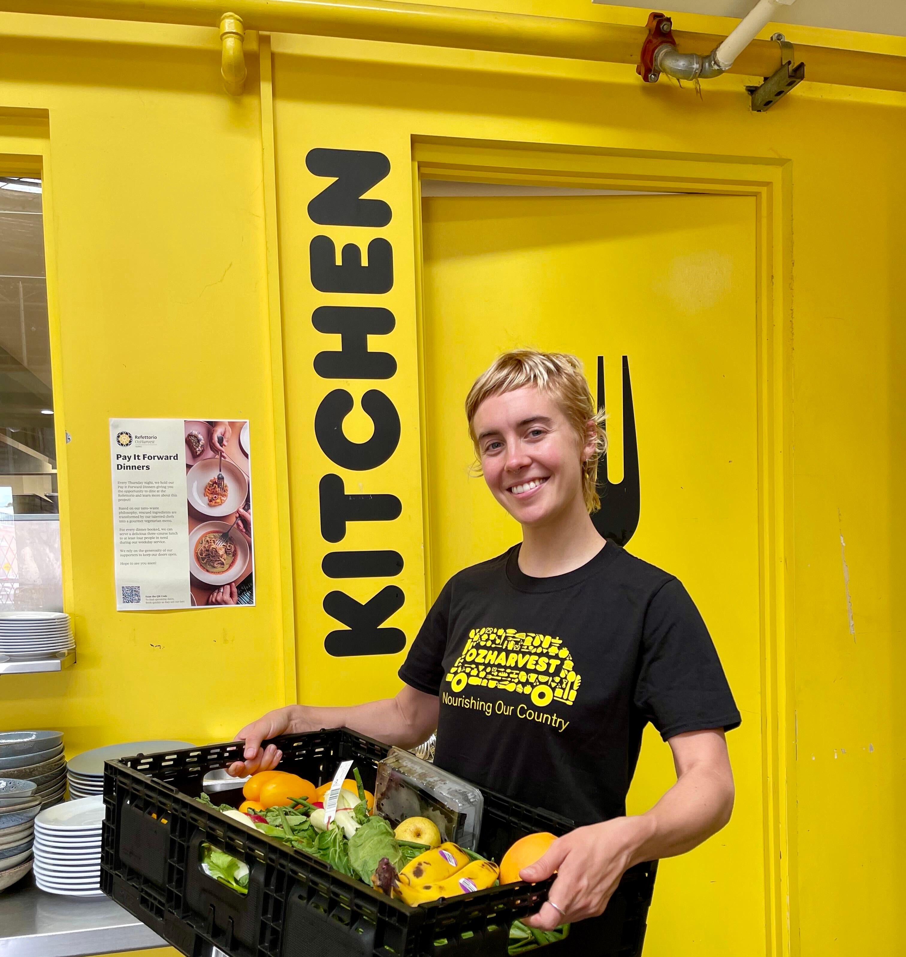 Ruby Wake at work in the OzHarvest kitchen, smiling and holding a box of vegetables.