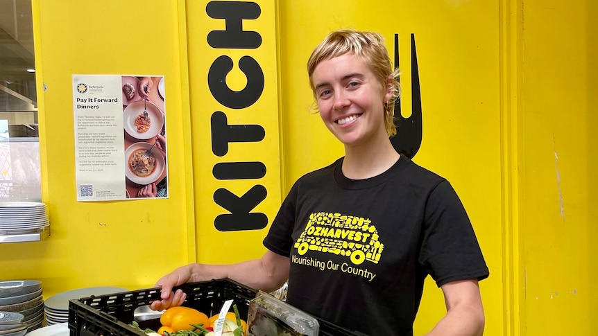 Ruby Wake at work in the OzHarvest kitchen, smiling and holding a box of vegetables.
