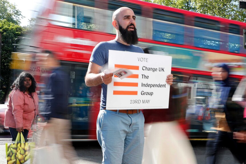 A bald man with a dark beard holds a Change UK party sign as a double decker red London bus whizzes past him.