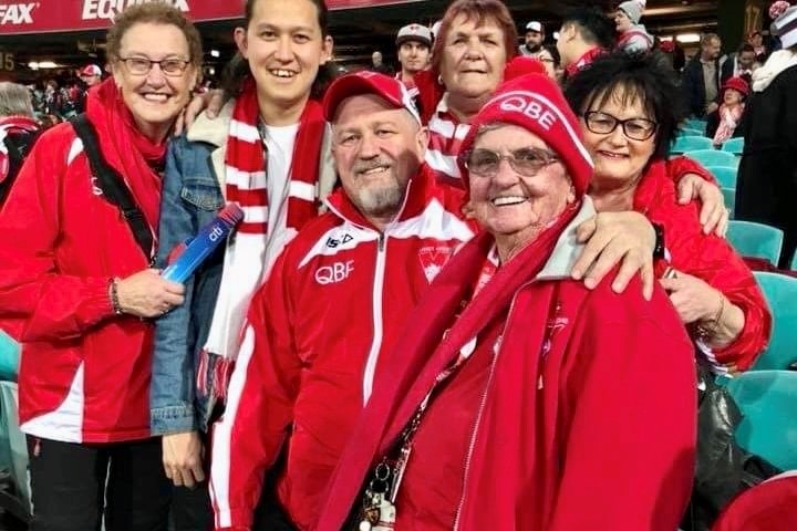 A group of six Sydney Swans fans dressed in club colors pose for a photo from the seats during a game