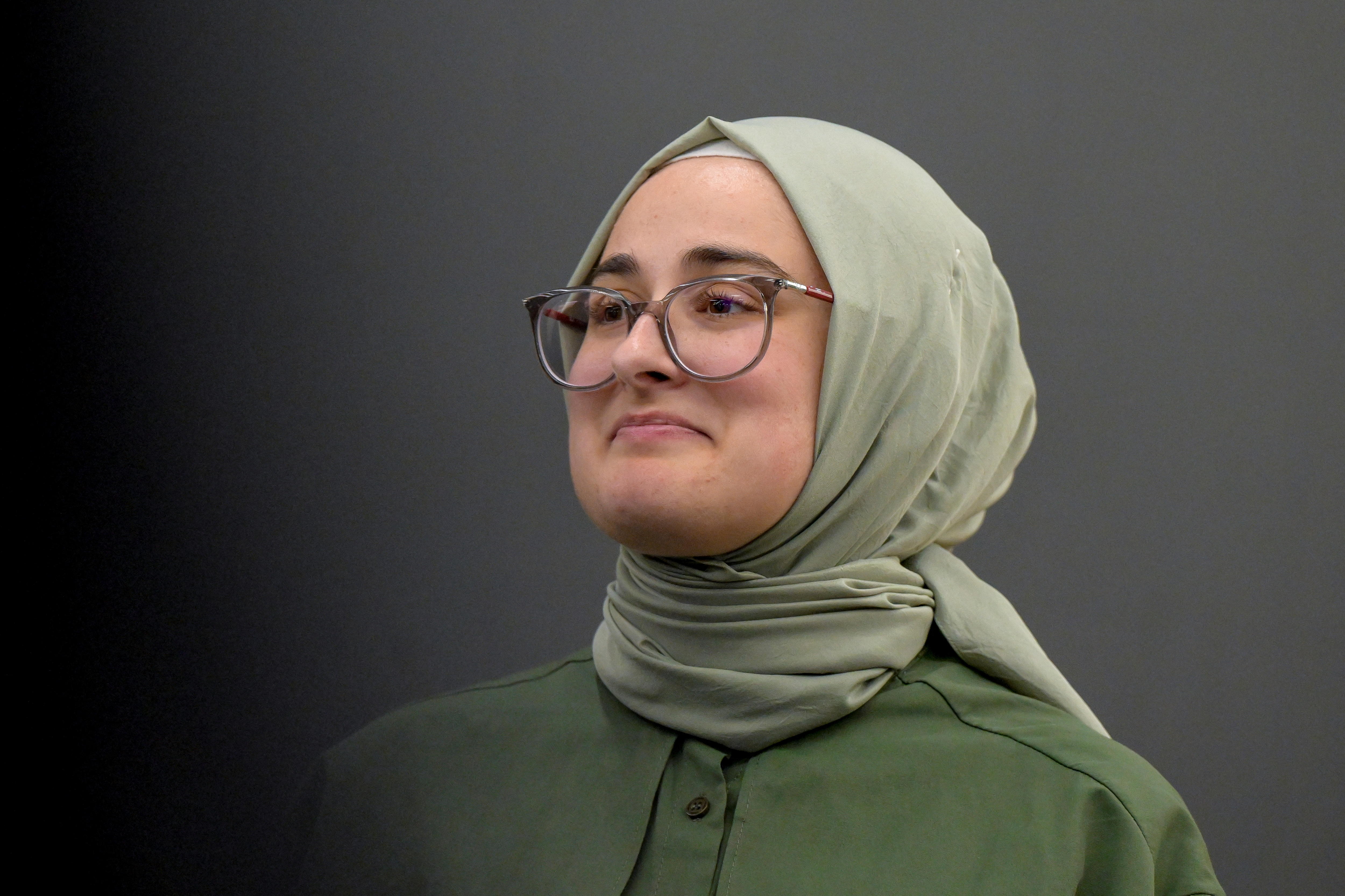 A woman wearing a green hijab and glasses smiles into the distance