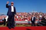 Trump standing in front of a rally of supporters with his fist raised, wearing a blue suit and red MAGA hat