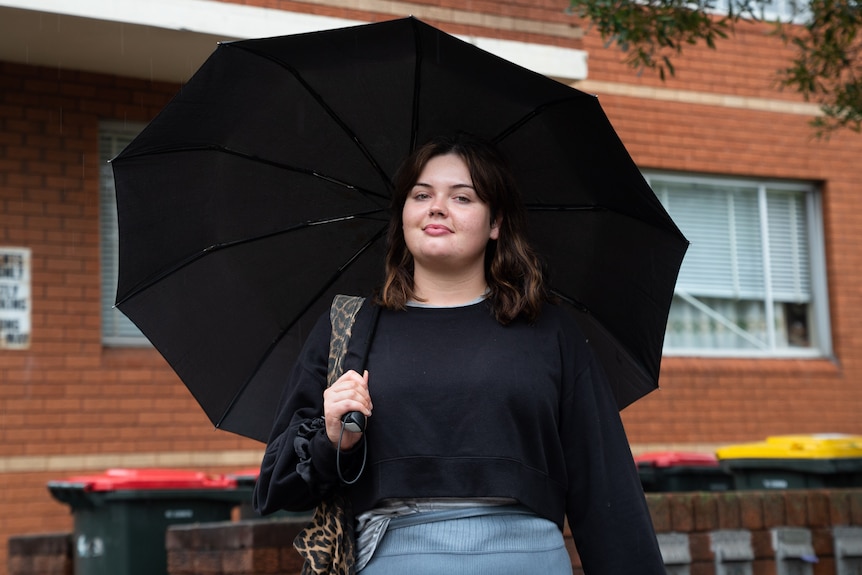 A woman holds a black umbrella, she's wearing a black long sleeve shirt in front of a red brick building and bins.