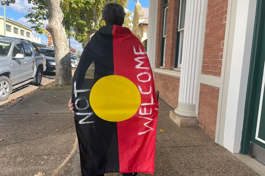 A woman wearing an Aboriginal flag as a cape that has the words 'not welcome' written on it.
