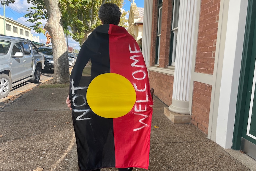A woman wearing an Aboriginal flag as a cape that has the words 'not welcome' written on it.