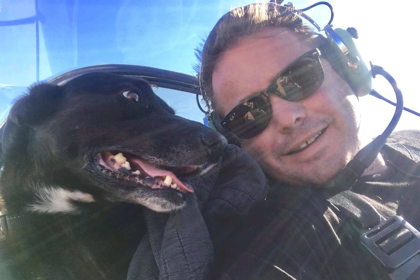 A man with headphones on in a plane with his black dog sitting next to him