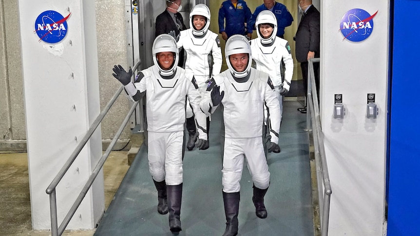 Four astronauts in white space suits walking down a ramp and waving