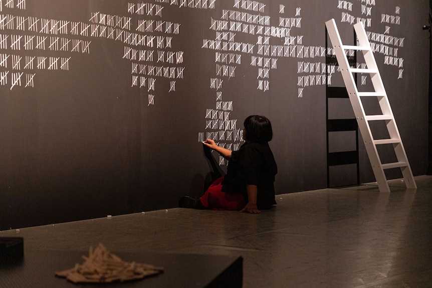A woman in a black shirt sits on the floor marking a tallty in chalk on the wall.