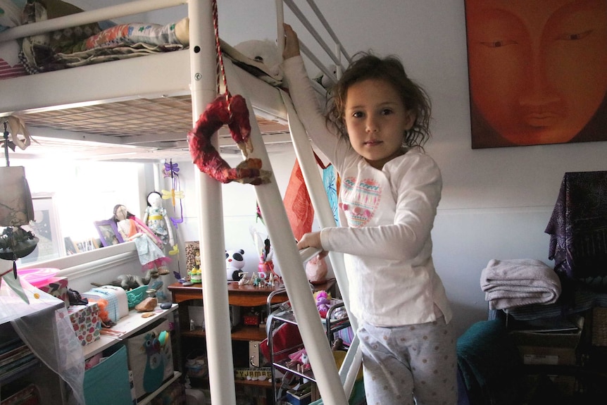 Four-year-old Zara stands on a ladder on her bunk bed in her bedroom looking at the camera.