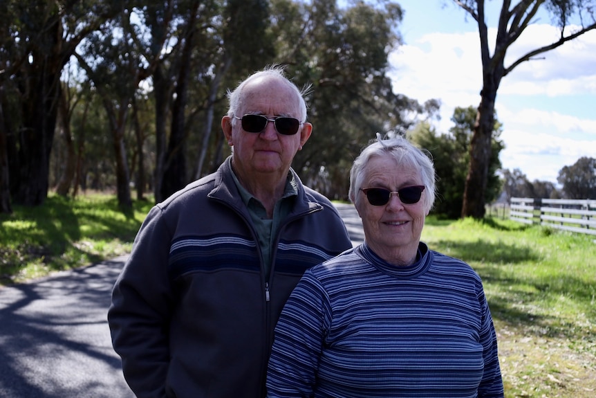 An elderly male and female couple, both wearing sunglasses, woman smiling a little, standing in front of a country road, trees. 