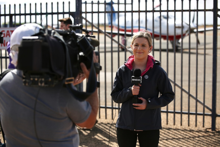 A woman with blonde hair holding an ABC microphone stands before a fence and a plane being filmed by a camera operator.