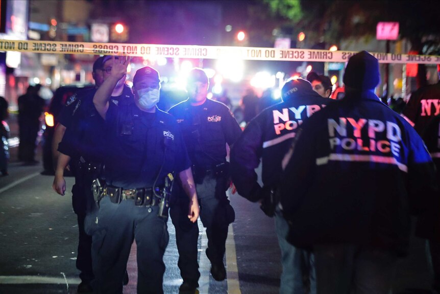 A police officer hold yellow police tape aloft to allow fellow officers to walk under as bright emergency lights shine.