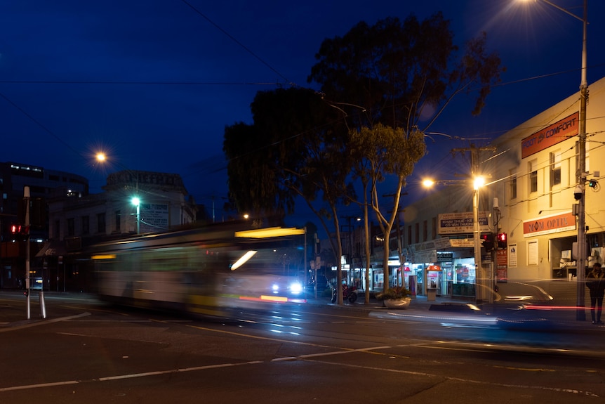 Against a dark sky, a blurry tram moves along a street, with a barely lit-up hotel front visible to the side.