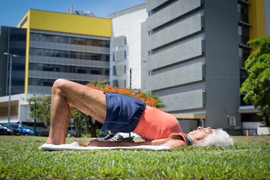 An elderly lady practices yoga poses in parklands in front of Cairns Hospital.