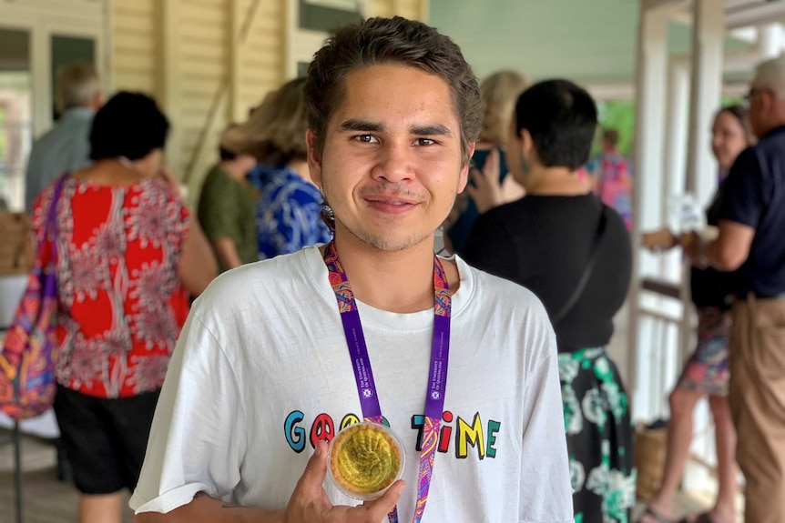 A young Indigenous man smiles, holding a wattle seed custard tart.