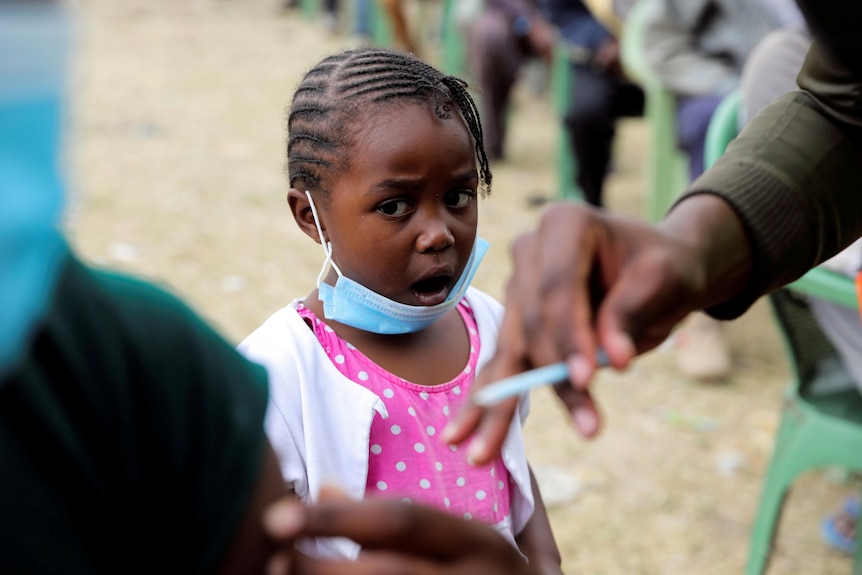 A little girl with a face mask pulled down stares with her mouth agape as a nurse holds a needle to a man's shoulder