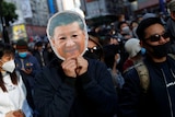 A person holds a paper mask depicting Xi Jinping's face up to their own, in a crowd of protesters
