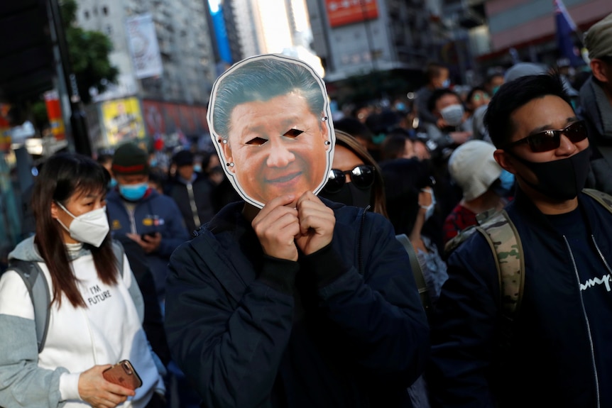 A person holds a paper mask depicting Xi Jinping's face up to their own, in a crowd of protesters