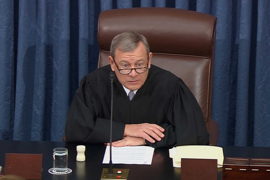A man in judge's robe sitting at a desk before a microphone looking annoyed