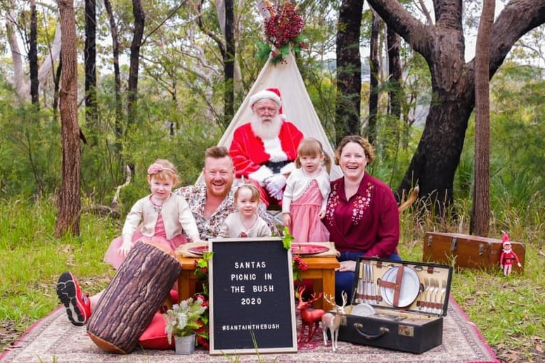A group of people sitting in front of a man dressed as Santa on a rug in the bush.