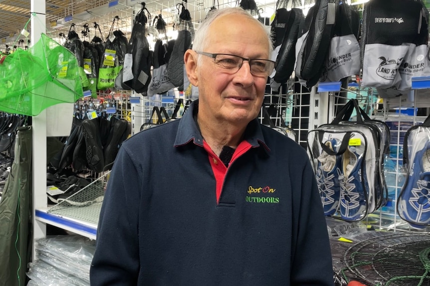 A smiling balding man, blue jumper with logo, glasses, grey hair looks off camera in a store filled with fishing gear.