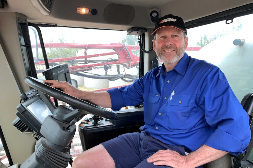 A man wears a blue shirt and cap, sitting behind the steering wheel of a large piece of farm machinery.