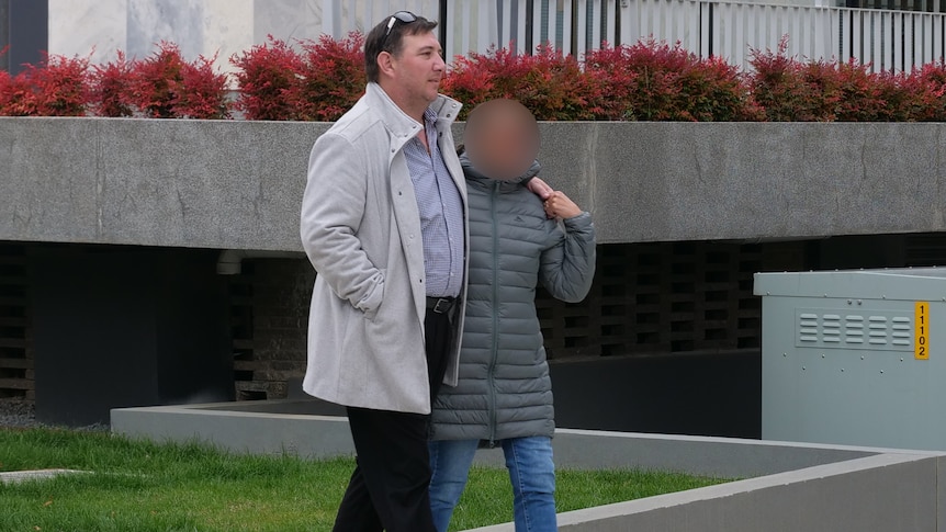 A man wearing a white jacket walks on the footpath with a woman around his left arm.