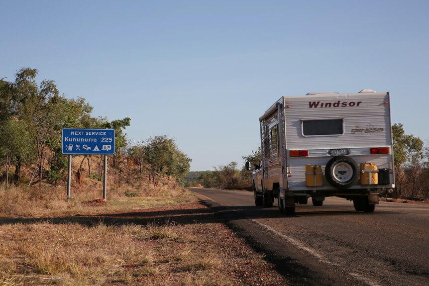 A motorhome moves along a remote stretch of highway in the NT, with a road sign visible.