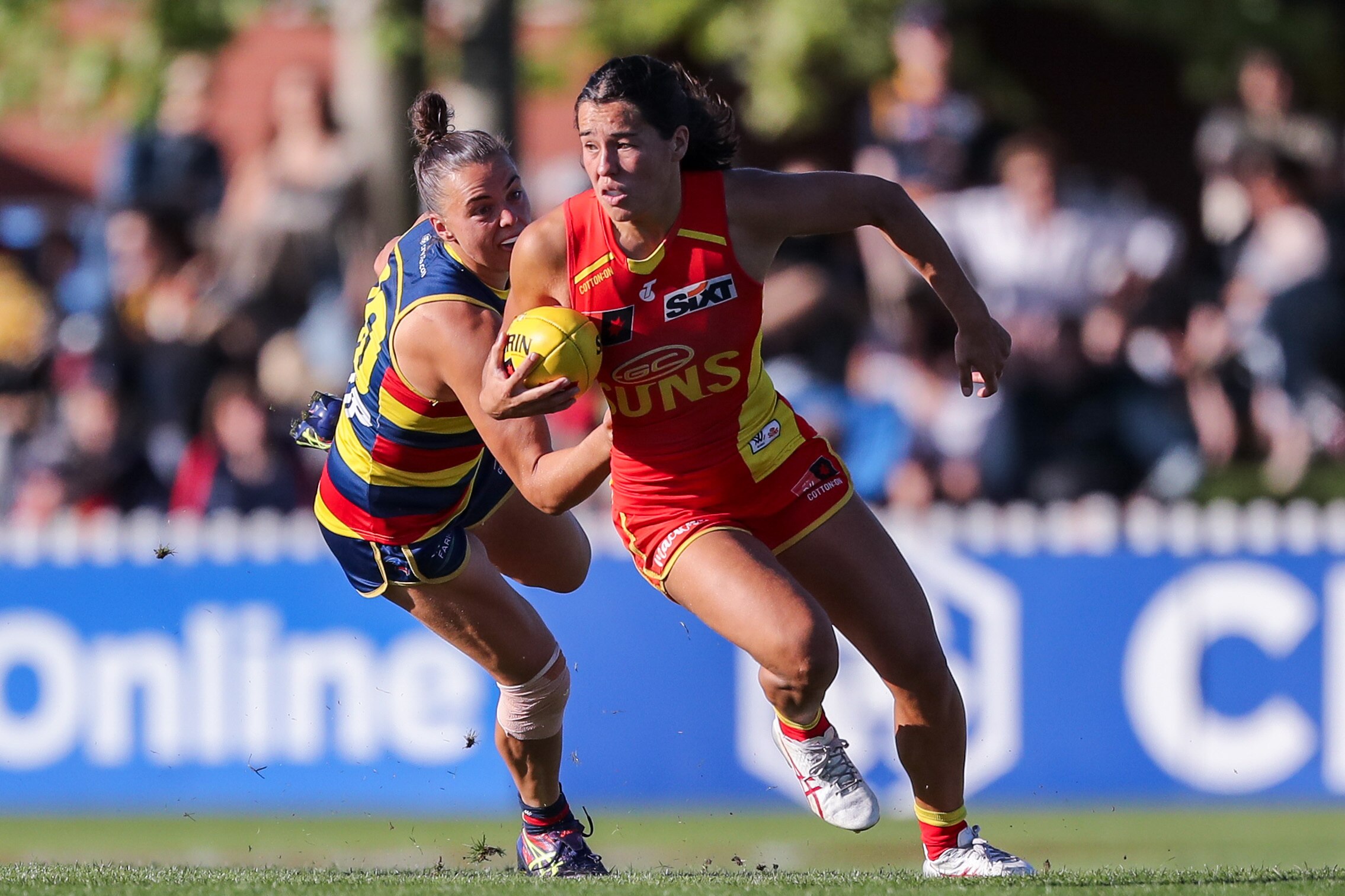 A Gold Coast Suns AFLW player holds the ball with her right hand as a Crows opponent attempts a tackle.