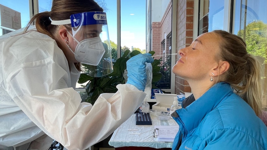 A healthcare worker in PPE swabs the nose of a woman wearing a blue jacket.