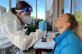 A healthcare worker in PPE swabs the nose of a woman wearing a blue jacket.