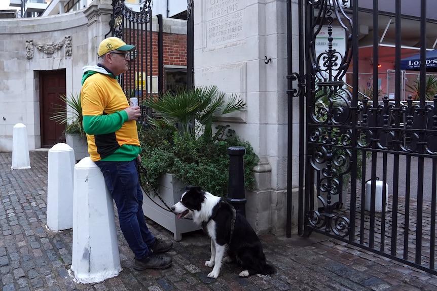 A man in green and gold Australian clothes stands outside Lord's after Shane Warne's death with his border collie dog.