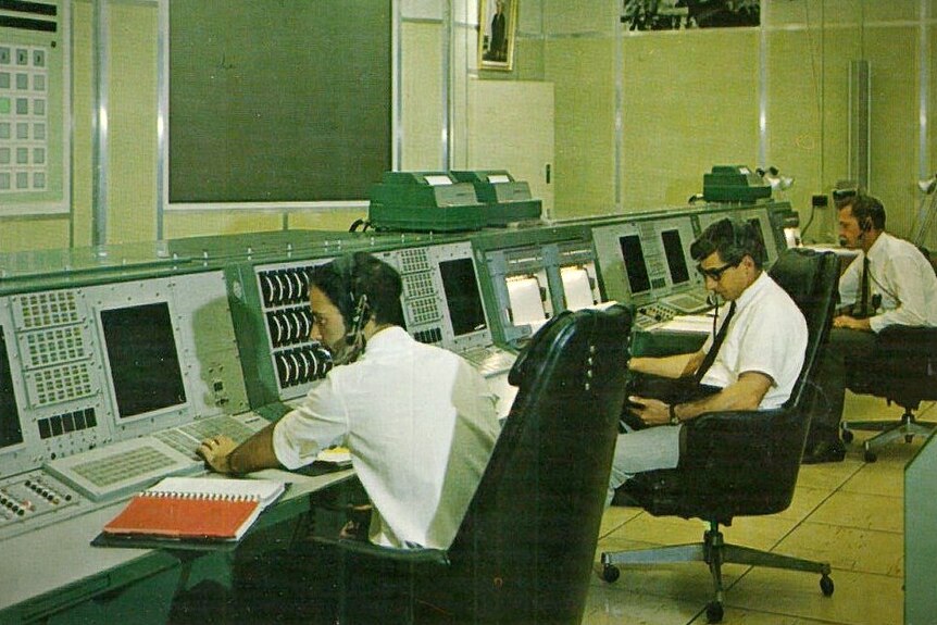 A wide shot of men in a communications room with controls and monitors.