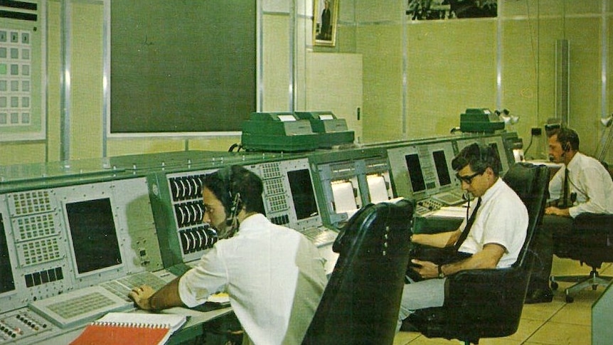 A wide shot of men in a communications room with controls and monitors.