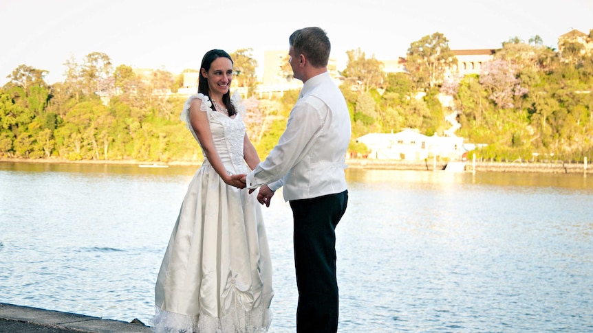 Marie and Damian Trebilco sit on a bench and smile in their wedding clothes