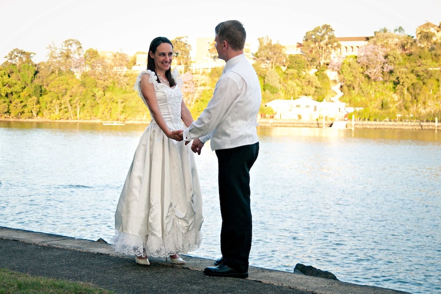 Marie and Damian Trebilco sit on a bench and smile in their wedding clothes