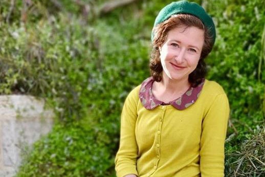 A woman wearing a green beret sits on a low wall in front of a wall of greenery.